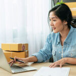 Woman sitting at a table typing on a laptop with boxes stacked up next to her and behind her on a shelf