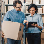 Man holding a box in a warehouse speaking to a woman holding an ipad
