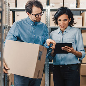 Man holding a box in a warehouse speaking to a woman holding an ipad