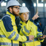 Portrait of Male and Female Industrial Engineers in Hard Hats Discuss New Project while Using Laptop. They Wear Safety Jackets.They Work at the Heavy Industry Manufacturing Factory.