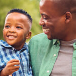 A close-up view of a 3-generation family, grandfather, father, and toddler, all with big smiles as the grandfather and father look inwards at the toddler.