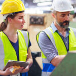 Male and female working in a plant looking at a machine wearing hard hats and a vests