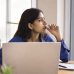 A woman sitting at a desk, with a computer and notepad in front of her, looking off to her left with a pondering expression on her face while biting her thumb nail.