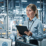 Car Factory Office Concept: Portrait of Successful Female Chief Automotive Engineer Using Tablet Computer to Design and Optimize Automated Robot Arm Assembly Line Manufacturing Electric Vehicles