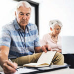 A older male and female sitting together while the male writes on a notepad with an agenda in his other hand and the female sits more in the distance typing on a computer.