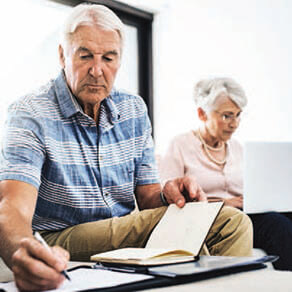 A older male and female sitting together while the male writes on a notepad with an agenda in his other hand and the female sits more in the distance typing on a computer.