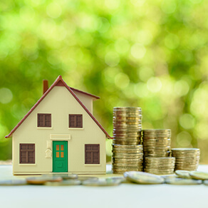 View of the front of a pretend house with five stacks of coins to the right of it and coins spread out in front and blurry green background
