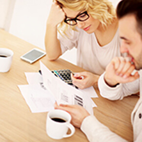 Blonde hair lady with black glasses sitting at table with man looking at their financial information, coffee cups, calculator and phone on table
