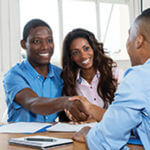 Two men at a desk shaking hands and a lady smiling at one of the guys