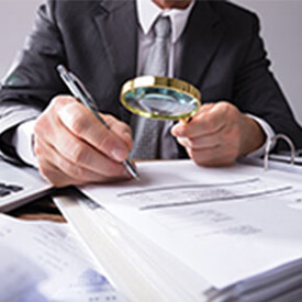 Man using magnifying glass to review papers, has a pen in hand