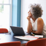 Lady sitting at a table with laptop, papers and glasses looking out a window in deep thoughts